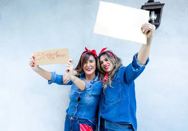 Two exuberant women take a selfie, one holding a sign 'Strong Women', celebrating their joy and empowerment