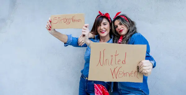 Two enthusiastic women share a moment of solidarity, taking a selfie with signs \'Strong Women\' and \'United Women\', embodying empowerment and unity.