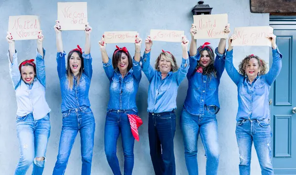 A group of exuberant women in denim, with red bandanas, cheerfully raise signs with messages of empowerment and solidarity.