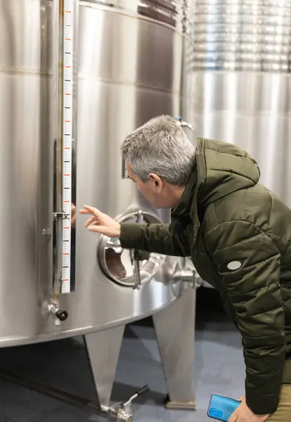 Winemaker attentively checking the level indicator on a stainless steel wine tank in a winery.