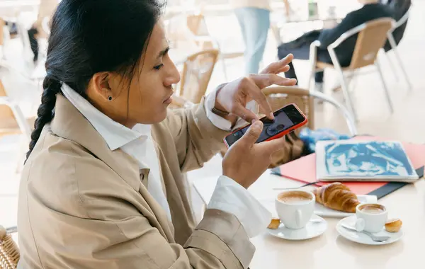 A focused young professional uses her smartphone at a cafe, surrounded by a fresh cup of coffee and pastries.