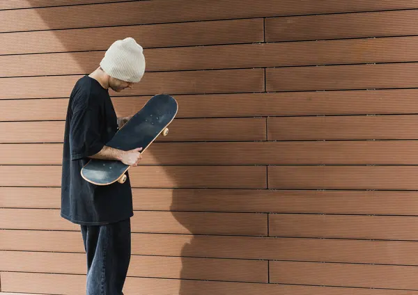 熟考中のスケートボーダーは テクスチャーされた木製の壁に対してスケートボードを保持し 都市の内観の感覚を呼び起こします — ストック写真