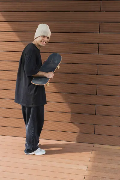 一个快乐的滑板运动员举着他的滑板 在木墙边对着摄像机微笑 流露出一个友善的都市氛围 — 图库照片
