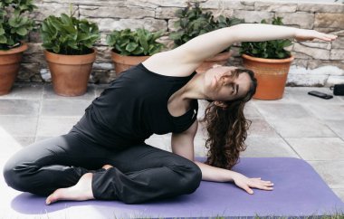 Odaklanmış bir kadın, saksı bitkileriyle dolu sakin bir bahçede yoga minderinde yan esneme hareketi yapıyor..