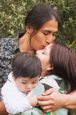 Ekvadorlu baba, bebeğini bir bahçede tutarken annesini alnından öper ve derin aile sevgisini ifade eder.