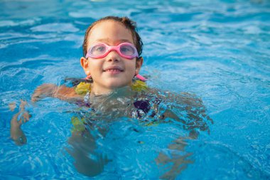 Pembe gözlüklü mutlu genç bir kızın yakın çekimi, açık mavi bir havuzda yüzerken gülümsüyor.