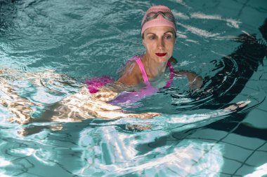 Bir dişi yüzücü, kısmen açık mavi suya batmış bir şekilde kapalı bir yüzme havuzunda zaman geçirmekten zevk alır. Pembe bir mayo ve yüzme şapkası giyiyor. Rahatlama ve memnuniyet hissini yansıtıyor..