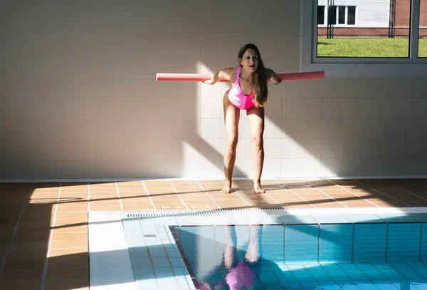 Pembe mayo giymiş atletik bir kadın yüzmeden önce havuzun güvertesinde havuz eriştesiyle ısınma egzersizleri yapıyor..