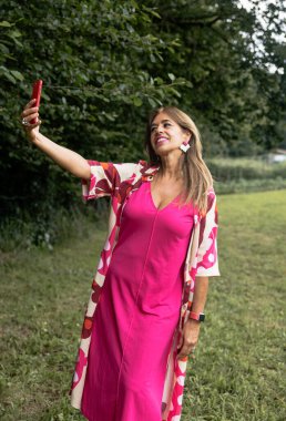 Canlı pembe elbiseli ve çiçekli ceketli olgun bir kadın yeşil kırsal bir ortamda selfie çekmek için poz veriyor, açık havanın tadını çıkarıyor..