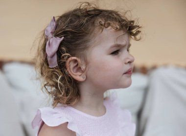 İki yaşında, kıvırcık saçlı, açık pembe elbisesi ve uygun bir yayı olan bir kızın yakın plan portresi..