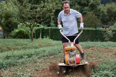 Açık mavi gömlekli bir adam sebze bahçesinde bir dümen işletiyor, toprağı ekime hazırlıyor. Görüntü, bahçecilik ve toprak ekimine odaklanmış açık hava çalışmalarını yakalıyor..