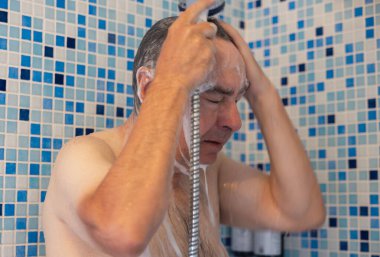 Orta yaşlı bir adam duş alırken ve saçını yıkarken görülüyor. Bir duş başlığı tutuyor ve kafasındaki sabunu temizliyor..