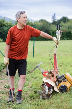 Küçük bir bahçe traktörünün yanında duran bir adam, dışarıda çiftçilik yapmaya hazırlanıyor..