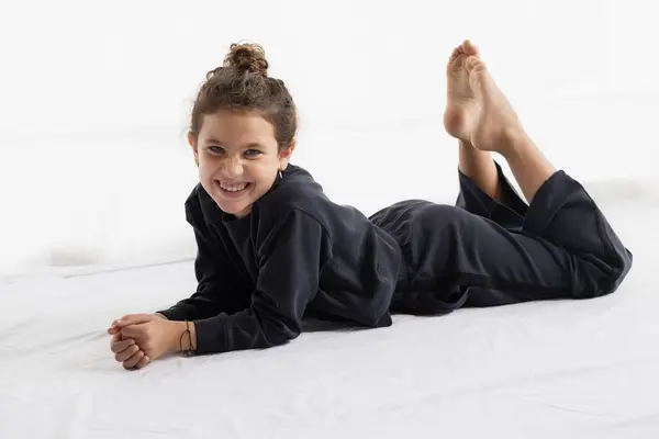 Neşeli genç bir kız beyaz bir yatakta uzanıyor, günlük koyu bir kıyafet giyiyor, profesyonel bir stüdyo ortamında parlak bir şekilde gülümsüyor. Reklamcılık ya da tanıtım kampanyaları için ideal. Neşe ve gençlik gösterisi..