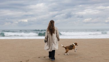 Genç kadın ve neşeli av köpeği kumlu bir sahilde, arka planda dalgalar var.