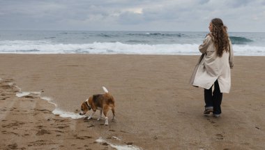 Bulutlu bir günde köpeğiyle birlikte okyanusun kenarında trençkotlu bir kadın.