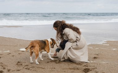 Genç bir kadın köpeğiyle oynamak için okyanusun kenarındaki kumsala çömeliyor..