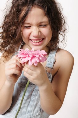 Kıvırcık saçlı, kot pantolonlu, elinde pembe bir çiçekle gülümseyen neşeli kız..