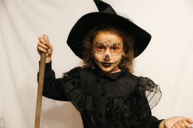 A young girl dressed as a witch with intricate skull face paint, holding a broom, posing indoors. clipart