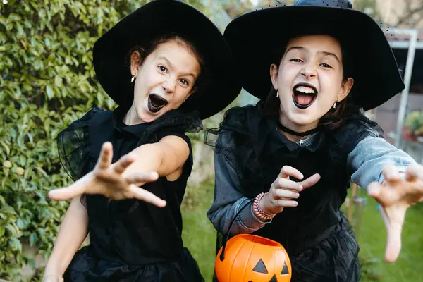 Cadı kostümlü iki heyecanlı kız kameraya bakıyor ve bahçede oynuyorlar..