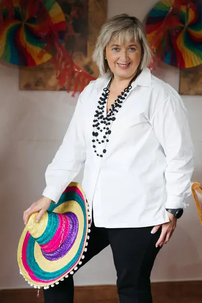 Beyazlar içinde şık giyinmiş, elinde canlı bir sombrero tutan ve kapalı alandaki şenlikli kutlamaya gülümseyen son sınıf öğrencisi bir kadın.