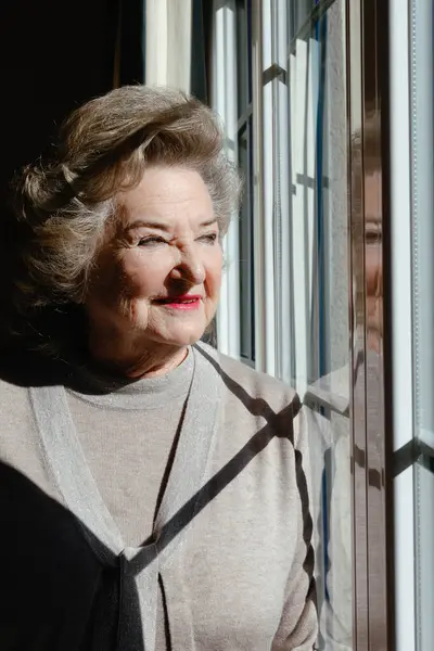 Gülümseyen son sınıf öğrencisi kadın yüzünde güneş ışığıyla pencereden dışarı bakıyor..