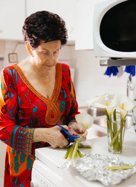 Mutfakta çiçek düzenleyen yaşlı kadın elinde makas ve cam vazo tutuyor.