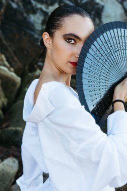 Güzel kız plaj kayalarının yanındaki yelpazeyle kameraya bakıyor.