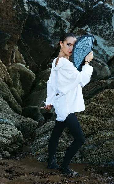 Flamenko tarzında kayalık sahilde yelpazeyle poz veren genç bir kadın.