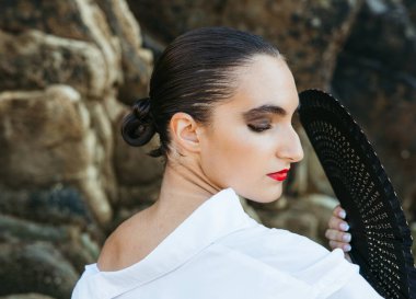 Dramatik makyajlı zarif bir genç kadın elinde yelpaze, gözleri kayalık sahil şeridinde kapalı poz veriyor.