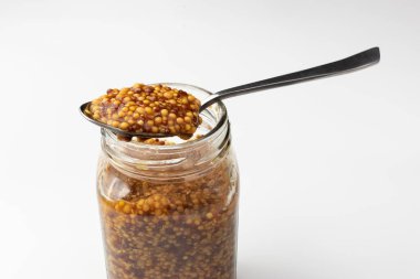 Kavanozda tahıllı hardal ve beyaz arka planda kaşık..