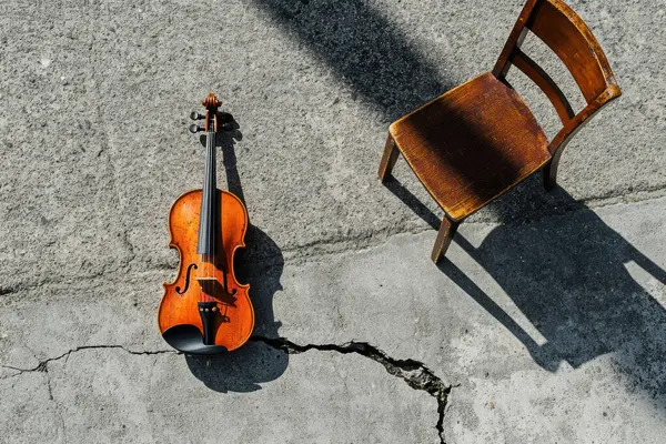 Boş ahşap bir sandalyenin yanında yerde yatan bir keman huzur ve tefekkür sahnesi yaratır. Güneş ışığı gölgeler oluşturur, anın duygusal derinliğini arttırır..