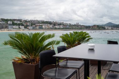 Bask bölgesindeki San Sebastian marinasında moda bir bar masası. Masada beyaz bir kül tablası ve iki deri koltuk var. Arka planda La Concha 'nın şehir plajını görebilirsiniz..