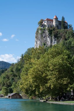 Sonbaharın sakin bir gününde göl kıyısından Kanlı Kale 'nin manzarası. Kale dağın zirvesinden şehre hükmeden kayalık bir uçurumun kenarında duruyor. Ahşap bir ev ve su yüzeyinde yürüyüş yapan insanlar görüyoruz..