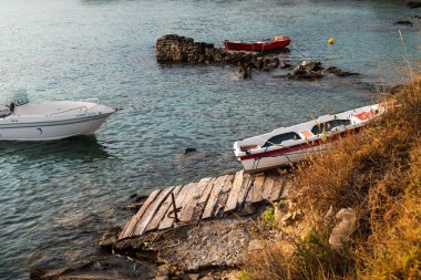 Romantic coastal view featuring three small boats anchored near rocky skerries: two are moored and one was hauled out of the water into dry land. clipart