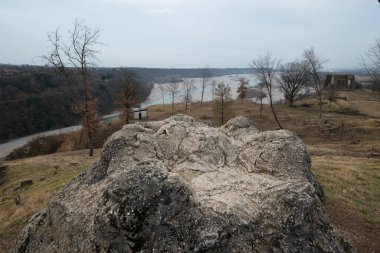 The germanic shrine of Pinzano and the Tagliamento river in Friuli (Italy) in a foggy morning of winter with a big rock in the foreground, bare trees and dry soil. The river is flowing in the background clipart