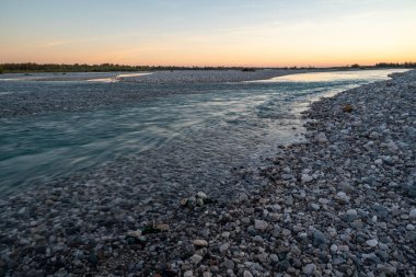 The Tagliamento river riverbed at dusk with, rocks and pebbles and sun rays reflections on the silky fresh waters in a long exposure effect clipart