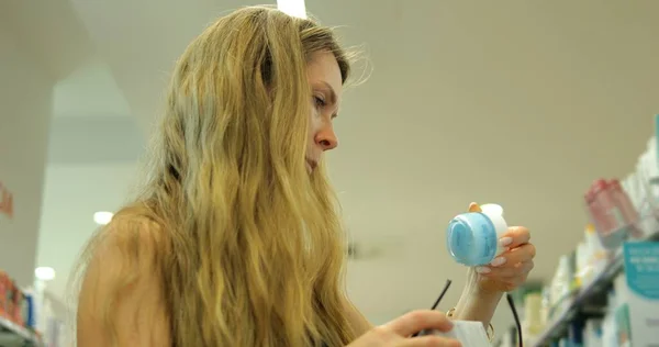 Düşük açılı genç kadın tüketici kozmetik mağazasında krem kavanozunu inceliyor. Eczanedeki güzel kadın, bilinçli ve satın alma kararını değerlendirmek için nemlendirici etiketini kontrol ediyor..
