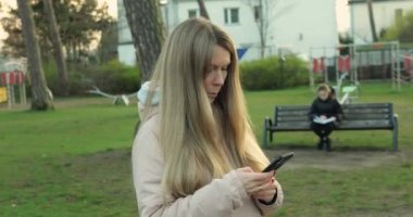 Parktaki çocuk parkında akıllı telefonuyla duran güzel bir kadın. Ceketli kadın, açık sonbahar öğleden sonra yeşil çimlerde cep telefonu kullanıyor..
