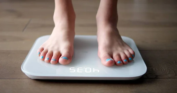 Young female teen stands barefoot on white scales to measure weight. Indicator value numbers show body mass, healthy dieting nutrition and fitness concept. High quality photo