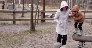 Anne oğluyla ahşap oyun parkında oynuyor ve yaylı kirişten kütüğe zıplamaya yardım ediyor. Beş yaşındaki çocuk, soğuk bir sonbahar gününde annesiyle dışarıda vakit geçirmekten hoşlanıyor..