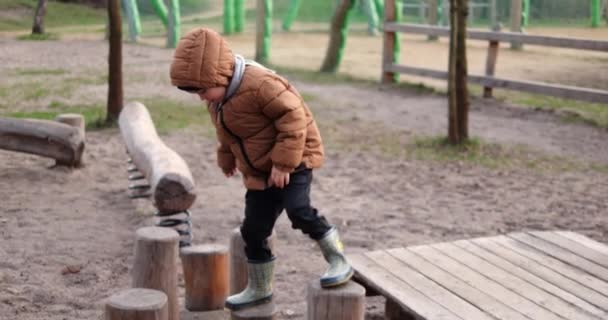 6歳の少年は 秋に木の遊び場にログをステッピングするように慎重に動きます 息子は 寒い日に遊具に挑戦的な障害を克服するママと屋外で余暇時間を過ごします — ストック動画