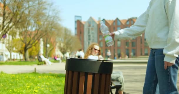 年轻的无脸女子把空塑料瓶扔进街上的垃圾箱 无名女孩的头发辫子用垃圾箱站在城市公园的长椅旁边 保持城市环境清洁的概念 — 图库视频影像
