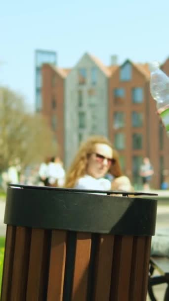 垂直ビデオ 匿名の女性は 空のプラスチックボトルを投げ 市立公園の通りでビンを無駄にします 髪のブレードを持つ未知の少女は ベンチの近くにゴミを使用しています 環境をきれいに保つコンセプト — ストック動画