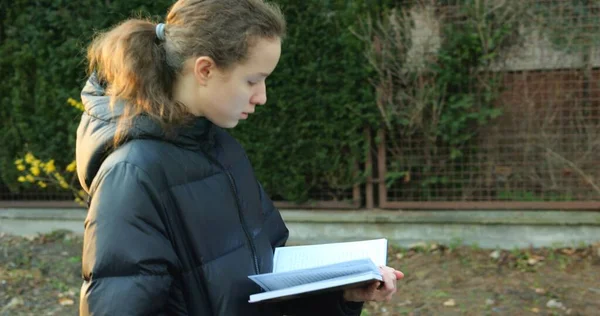 Sokakta yürüyen ve kitap okuyan gençlerin orta açıdan görünüşü. Okullu kızın, sonbahar günü gezinirken kitap okumaya odaklanmış bir fotoğrafını çekiyorum..