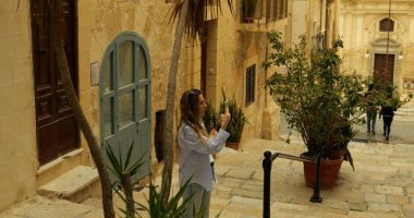 Turist kolej öğrencisi eski şehir merkezindeki tarihi simgelerin fotoğrafını çekiyor. Sıcak bir yaz günü Malta 'nın Valletta kentindeki ortaçağ binaları arasında cep telefonlu bir kadın. 4k görüntü