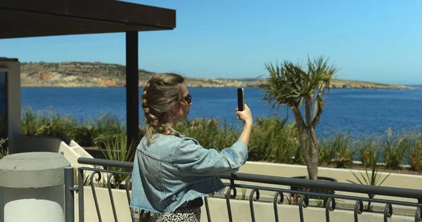 Young stylish woman shoots video of sea area landscape on smartphone at seafront. Middle age female traveler in denim jacket records story reel on sunny day.