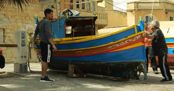 Marsaxlokk, Malta - 1 Mayıs 2023 - Maltalı balıkçı grubu Marsaxlokk köyünün eski caddesindeki otantik Malta teknesini elle taşımaya çalıştı.