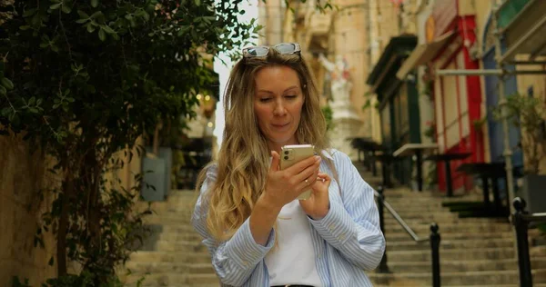 Young woman in sun glasses in city center old town swipe smartphone enjoy photo. Female adult on old stairs in downtown on summer day smile checks instant messages in social media on phone. 4k footage