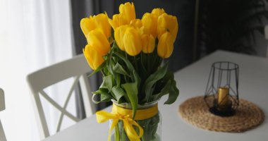 Pencerenin yanındaki büyük yemek masasında güzel sarı laleler. Süslü cam vazodaki yoğun renkli çiçekler yaz atmosferini oluşturur. Yüksek kalite fotoğraf